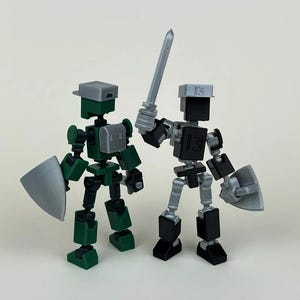 May include: Two small, articulated robot figures. One is green with a gray helmet and shield, the other is black with a gray helmet and sword. Both figures have posable arms and legs.