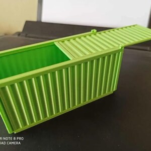 May include: A green plastic toy shipping container with a hinged lid. The container is open and the lid is propped up.