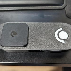 May include: A dark gray, rectangular device with a textured surface. It features a square section on the left with a circular logo and an oval section on the right with a white circular logo. The device is on a black surface.