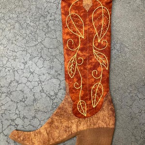 May include: A decorative fabric cowboy boot in shades of orange and brown. The boot has an embroidered leaf design in gold thread. A beige loop is attached at the top. The boot is designed to be a stocking.