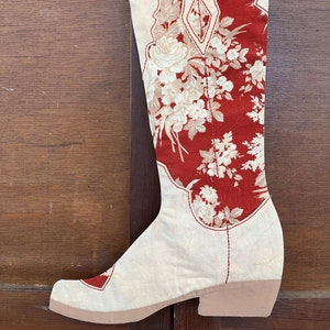 Puede incluir: Una bota de vaquero blanca de punto con un patrón floral rojo. La bota está decorada con costuras blancas y tiene un tacón rosa.