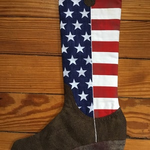 May include: A brown cowboy boot stocking with an American flag design. The boot is made of fabric and has a red, white, and blue pattern.