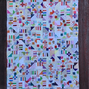 May include: A colourful patchwork quilt with a white background and a variety of geometric shapes and patterns. The quilt is made up of many different fabrics, including floral prints, stripes, and solids. The quilt is hanging on a wooden wall.