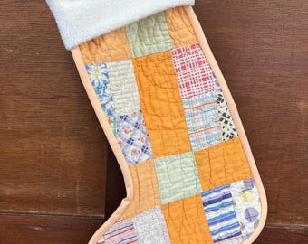 May include: A handmade Christmas stocking with a patchwork design. The body is a vibrant orange, composed of various fabric squares in different patterns and colors. The cuff is white, fluffy material, and the edges are trimmed with a light peach fabric.
