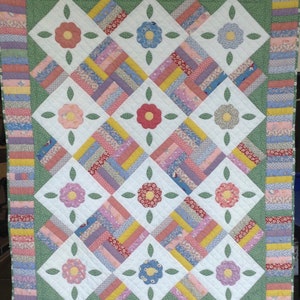 May include: A white quilt with a green border and a pattern of pink, yellow, and blue flowers. The quilt is made of patchwork fabric and has a diamond pattern.