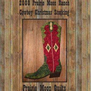 Może przedstawiać: Czerwono-zielona choinkowa skarpeta w kształcie kowbojskiego buta z diamentowym wzorem na cholewce. But znajduje się na drewnianym tle z tekstem "2008 Prairie Moon Ranch Cowboy Christmas Stocking" i "Prairie Moon Quilts".