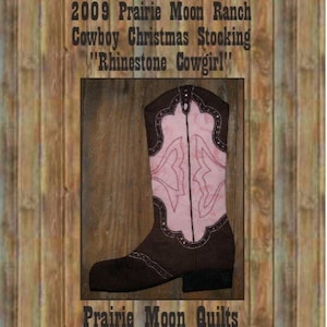 Pode incluir: Uma bota de cowboy marrom com costuras rosa e strass. A bota é uma meia de Natal com o texto "2009 Prairie Moon Ranch Cowboy Christmas Stocking "Rhinestone Cowgirl!" e "Prairie Moon Quilts".