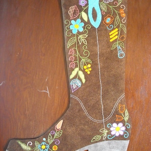 Pode incluir: Uma bota de couro marrom com uma fita azul e bordado floral colorido. A bota é decorada com flores em tons de laranja, amarelo, rosa, azul e verde.