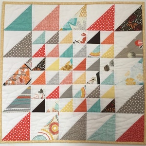May include: A square quilt with a patchwork design. The quilt features white squares with colorful triangles in teal, red, gray, yellow, and orange. The triangles have various patterns, including polka dots and floral prints. The quilt is bordered with a gold fabric.