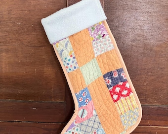 May include: A handmade Christmas stocking with a patchwork design. The stocking features a peach-colored body with various fabric squares in floral and patterned designs. The top is white, and it hangs from a wooden door.