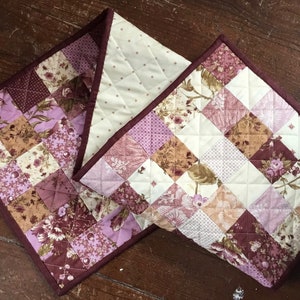 May include: A quilted table runner with a patchwork design featuring various shades of pink, brown, and white floral patterns. The runner has a burgundy border.