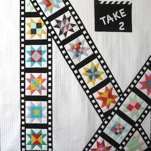 May include: A white quilt with two black and white film strips running diagonally across the fabric. The film strips are decorated with colorful patchwork squares. A black and white clapperboard with the text "TAKE 2" is in the upper right corner.