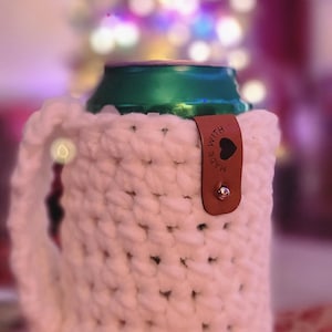 May include: A white, chunky-knit crochet can cozy with a handle, holding a green can. A small brown leather tag with a heart and the words "MADE WITH" is attached. The background is blurred with Christmas lights.