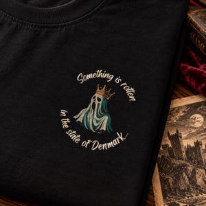 Könnte beinhalten: Ein schwarzes T-Shirt mit einer Grafik eines Geistes, der eine goldene Krone trägt. Der Text "Something is rotten in the state of Denmark..." ist in einem hellen Cremeton kreisförmig um den Geist herum aufgedruckt.