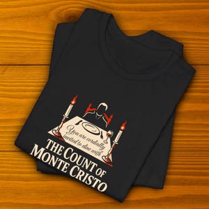 May include: Black t-shirt with a graphic design. The design features the text "You are cordially invited to dine with The Count of Monte Cristo." The graphic includes a dining table, candles, and a wine glass.