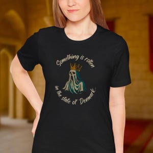 Könnte beinhalten: Schwarzes T-Shirt mit einer Grafik eines Geistes mit Krone und dem Text "Something is rotten in the state of Denmark." Das Design ist in Teal, Gold und Weiß gehalten. Das T-Shirt hat einen Rundhalsausschnitt.