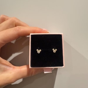 May include: A pair of gold-coloured stud earrings shaped like a mouse head, displayed in a pink and black jewellery box. The earrings are small and feature three round, sparkling accents.