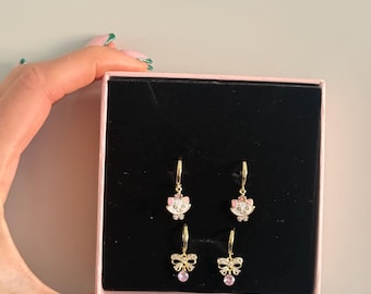 Puede incluir: Una caja rosa contiene dos pares de pendientes de aro dorados. Un par presenta una cara de gato blanco con detalles rosas, el otro un lazo con una gema rosa. Los pendientes se muestran sobre un fondo negro.