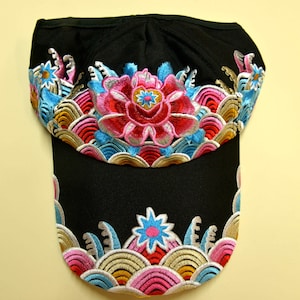 May include: Black baseball cap adorned with intricate floral embroidery. The design showcases a central pink flower with blue and yellow details, set against a backdrop of colourful wave patterns in blue, pink, and gold. The embroidery provides a decorative element.