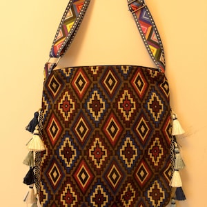 May include: A rectangular shoulder bag with a geometric diamond pattern in brown, red, and blue. The bag has a colorful patterned strap and is adorned with tassels in various shades of blue and white. The bag is approximately 12 inches wide and 14 inches tall.