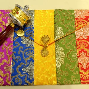 May include: A colorful prayer wheel and a decorative envelope. The prayer wheel is silver and gold with a wooden handle. The envelope is made of fabric in shades of purple, blue, yellow, green, and red, with gold floral patterns.