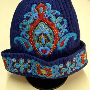 Puede incluir: Un gorro de punto azul oscuro con un diseño bordado central prominente en tonos turquesa brillante, rojo y amarillo. El borde doblado está decorado con una banda continua de bordados florales y de estilo cachemir en colores a juego.