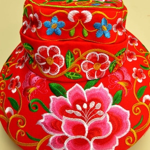 Pode incluir: Um chapéu vermelho vibrante com bordados florais elaborados. Apresenta grandes flores rosa e brancas, flores vermelhas e brancas menores e flores azuis, todas delineadas com fio verde e dourado. A aba e a copa são totalmente bordadas.