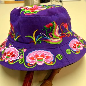 May include: A purple bucket hat featuring vibrant, multi-colored floral and bird embroidery. The design includes pink and red lotus-style flowers, green swirling vines, and a stylized bird in shades of pink, green, and yellow across the crown.