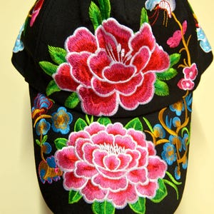 Puede incluir: Gorra de béisbol negra con bordados florales vibrantes. La gorra presenta grandes flores bordadas detalladas en tonos de rosa y rojo, con hojas verdes y acentos azules y amarillos. Un accesorio elegante.