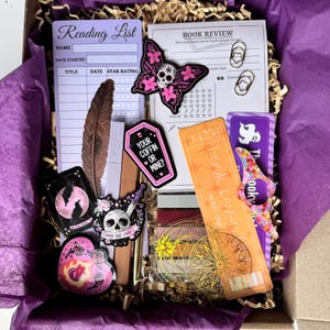 May include: A gift box containing stationery and decorative items. Features a reading list, book review sheet, stickers with skull and butterfly designs, a feather, bookmarks, and a ticket-shaped item. The box is lined with purple tissue paper.