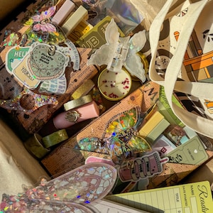 May include: A cardboard box filled with stationery and accessories including a canvas tote bag, a butterfly hair clip, a small candle with dried petals, various holographic stickers, a reading list card, and several small rectangular boxes.