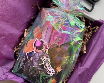 Op de afbeelding: Een geschenkdoos verpakt in iriserend cellofaan, vastgebonden met touw en genesteld in paars vloeipapier. De doos bevat stickers, een hanger en andere kleine items. De sticker zegt "I rather reading". Een schaar is zichtbaar op de achtergrond.
