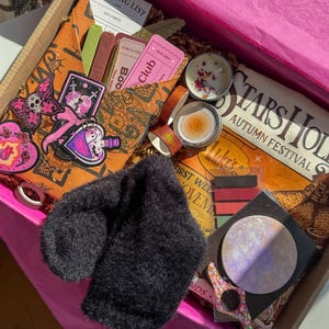 May include: A gift box filled with assorted items including a black fuzzy accessory, a Stars Hollow Autumn Festival sign, two small candles, a reading list card, a pink ticket, stickers featuring a skull butterfly and potion, and a moon-themed card.
