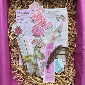 Puede incluir: Una caja rosa llena de artículos de papelería, que incluyen una lista de lectura, una hoja de revisión de libros, pegatinas, adornos en forma de mariposa, una pluma y un bolígrafo dorado. Las pegatinas tienen frases como "Read More Books".
