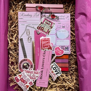 May include: A pink book lovers gift set with a reading list notepad, stickers, bookmarks, a pen, and a page holder. The set includes stickers with phrases like "Smutty Book Club" and "Enemies to Lovers". The set is in a brown box with pink tissue paper.