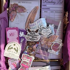 May include: A purple gift box containing stationery and stickers. The contents include a book review card, a reading list, and stickers with phrases such as "Trigger Warnings" and "My Book Boyfriends Have Wings".