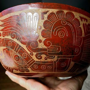 May include: A hand-held, red-toned bowl with intricate, carved designs. The artwork features stylized figures and patterns in a lighter, contrasting color, creating a striking visual contrast. The bowl's surface has a glossy finish.
