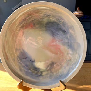 May include: A large, round ceramic serving bowl with a white glaze and a swirling pattern of blue, pink, and green. The bowl is on a wooden stand.