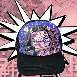 Pode incluir: Boné de caminhoneiro preto e azul com um design colorido de personagem de anime. O boné tem uma aba preta e uma parte traseira de malha, com uma ilustração vibrante no painel frontal. O design inclui um personagem com chifres rosa e um bocal de bambu.