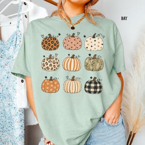 May include: A light sage green short-sleeve t-shirt featuring a grid of nine pumpkins with various patterns including leopard print, polka dots, stripes, floral, and plaid, accented with small black hearts and stars printed on the front.