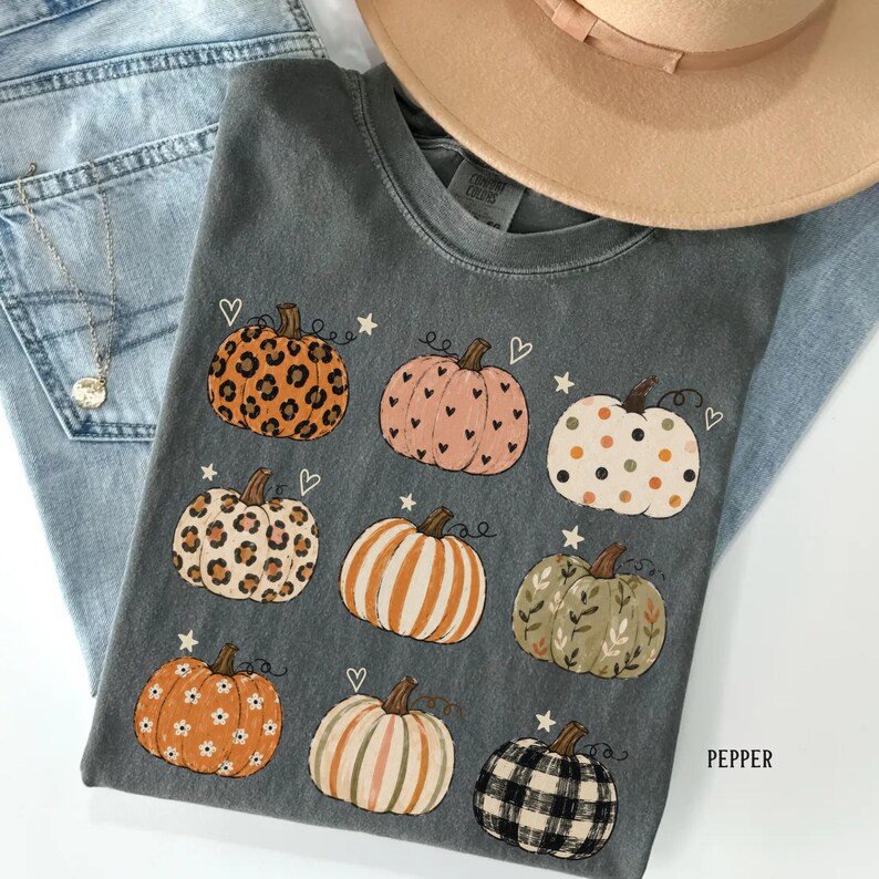 May include: A charcoal gray t-shirt featuring a graphic print of nine pumpkins with various patterns including leopard print, polka dots, stripes, floral motifs, and plaid, accented with small white hearts and stars across the fabric.