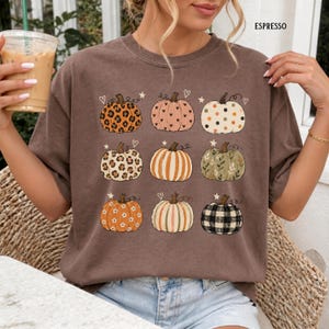 May include: A brown t-shirt featuring a graphic print of nine pumpkins in various patterns including leopard print, polka dots, stripes, floral, and plaid, accented with small hearts and stars, with the word ESPRESSO printed in the top right corner.