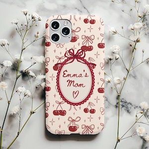 Puede incluir: Una funda de teléfono con fondo crema y un patrón repetido de cerezas, lazos y estrellas en color rojo intenso. El centro de la funda presenta un diseño ovalado con el texto "Emma's Mom" y un corazón.