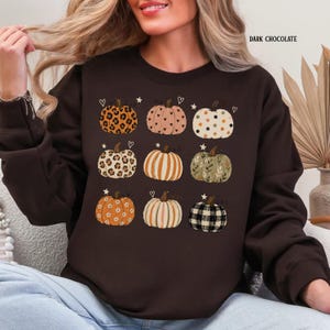May include: A dark chocolate brown long-sleeve sweatshirt featuring a graphic print of nine pumpkins in various patterns including leopard print, hearts, polka dots, stripes, floral, and plaid, with small stars and hearts scattered around them.
