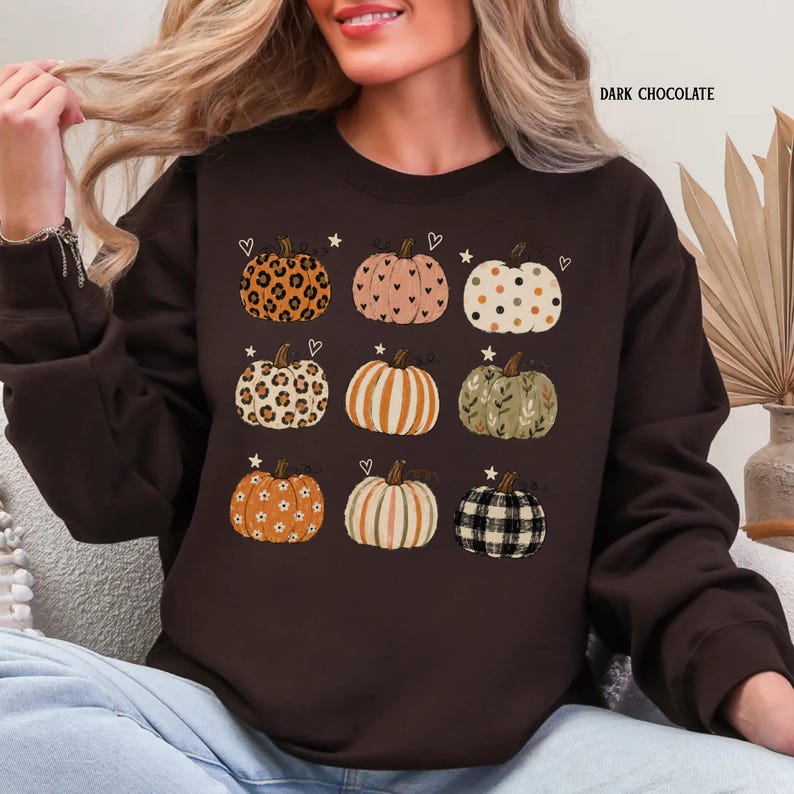 May include: A dark chocolate brown long-sleeve sweatshirt featuring a graphic print of nine pumpkins in various patterns including leopard print, hearts, polka dots, stripes, floral, and plaid, with small stars and hearts scattered around them.