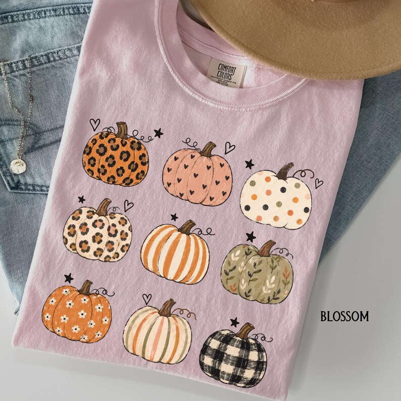 May include: A light pink t-shirt featuring a graphic print of nine pumpkins with various patterns including leopard print, polka dots, stripes, floral, and plaid, accented with small hearts and stars. The inner neck label reads Comfort Colors.