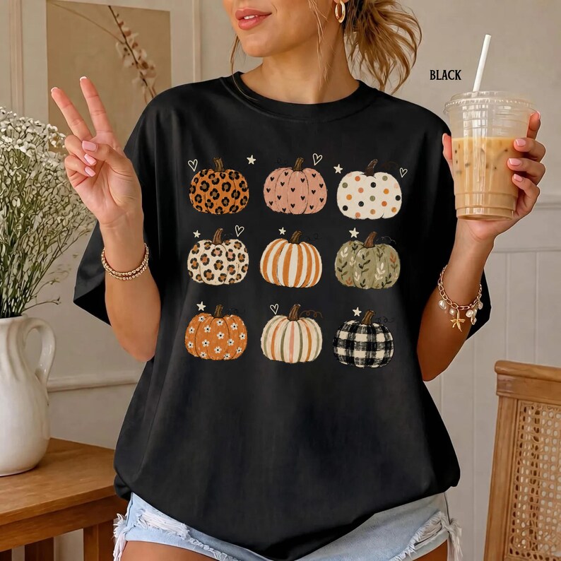 May include: A black short-sleeve t-shirt featuring a graphic print of nine pumpkins in various patterns, including leopard print, polka dots, stripes, and plaid. Small white heart and star icons are scattered between the pumpkins on the front.