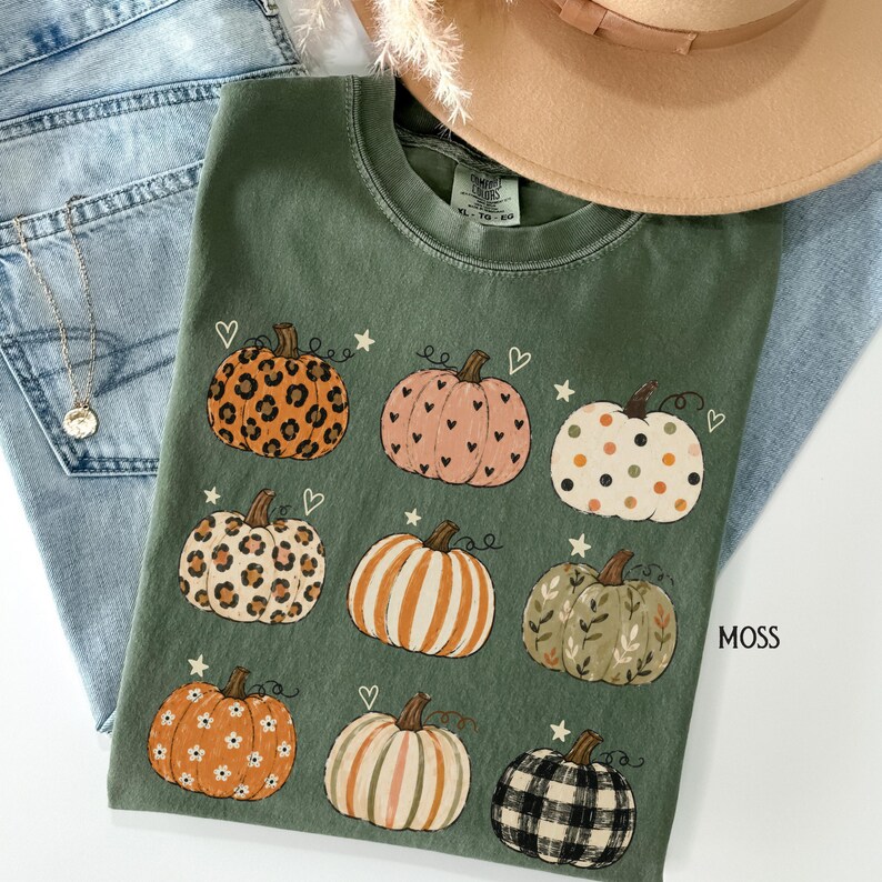 May include: A moss green t-shirt featuring a graphic print of nine pumpkins with various patterns including leopard print, polka dots, stripes, floral, and plaid, accented with small hearts and stars. The shirt has a crew neckline and a size XL tag.