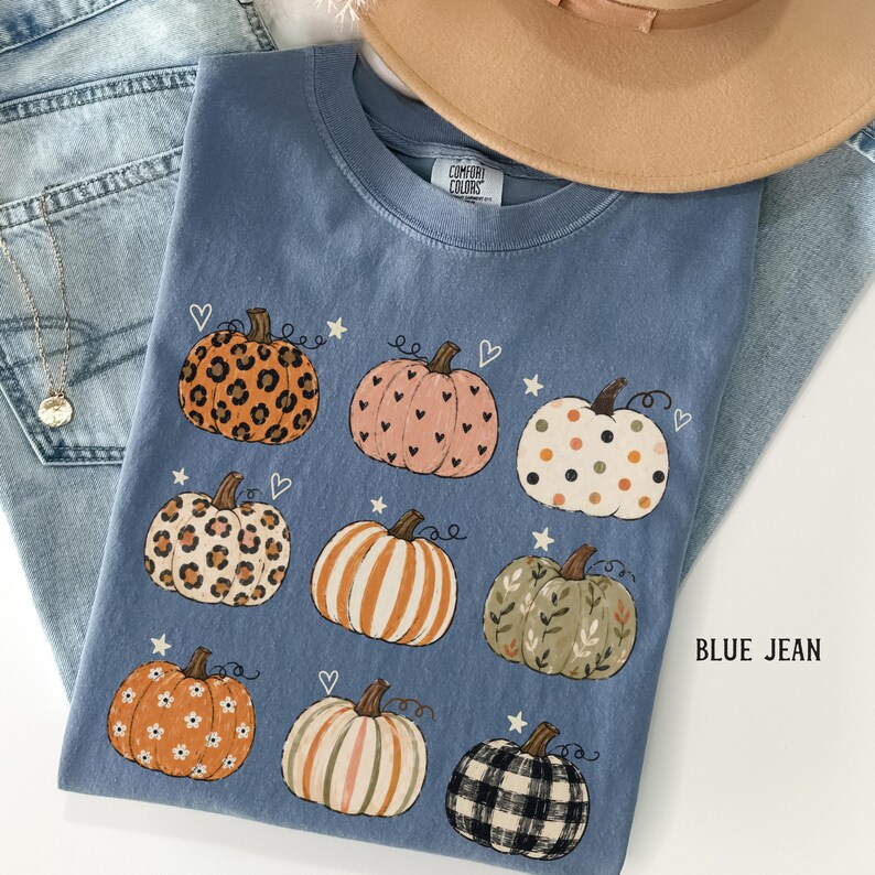 May include: A blue crew neck t-shirt featuring a graphic print of nine pumpkins with various patterns including leopard print, polka dots, stripes, floral, and plaid, accented with small white hearts and stars. The shirt has a Comfort Colors label.