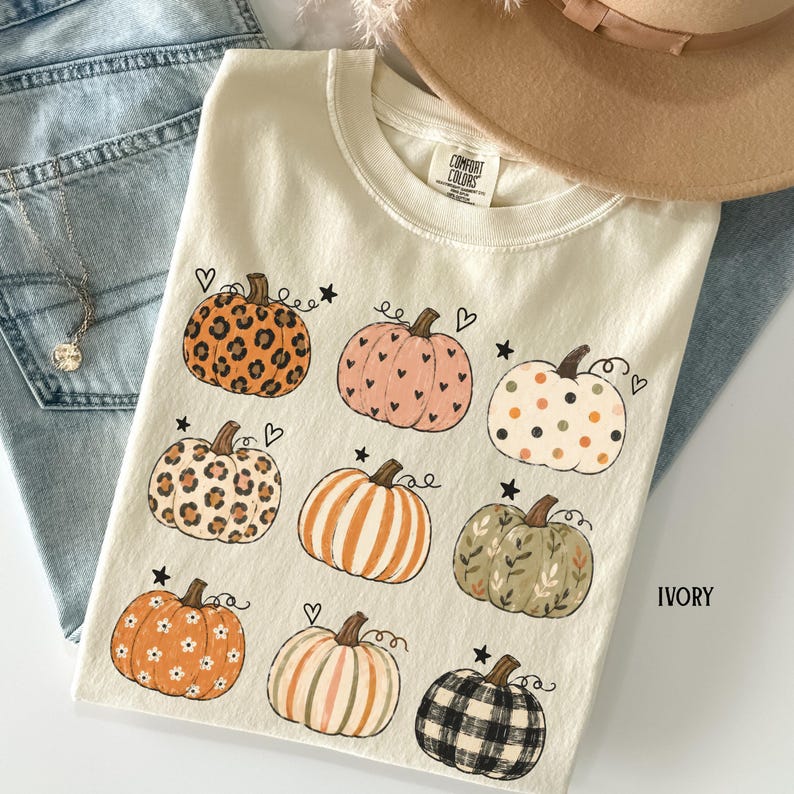 May include: An ivory short-sleeve t-shirt featuring a graphic print of nine pumpkins with various patterns including leopard print, polka dots, stripes, floral, and plaid, accented with small hearts and stars. The neck label reads Comfort Colors.